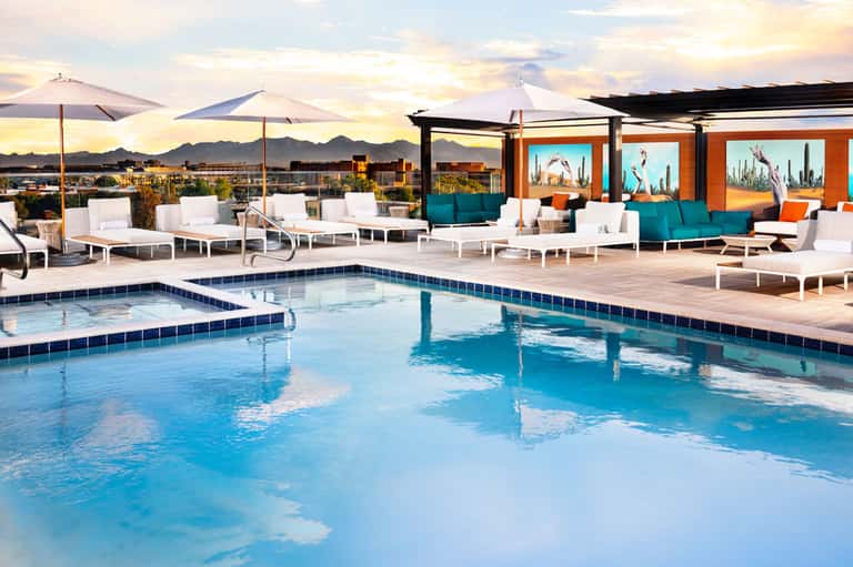 Resort pool deck at sunset with lounge chairs, umbrellas, and colorful modern buildings in background