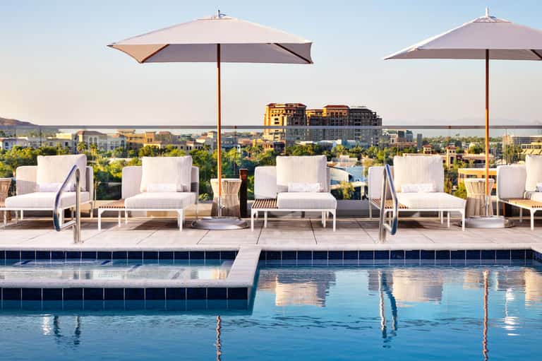 Resort rooftop pool deck with white loungers, umbrellas, and city skyline views at sunset