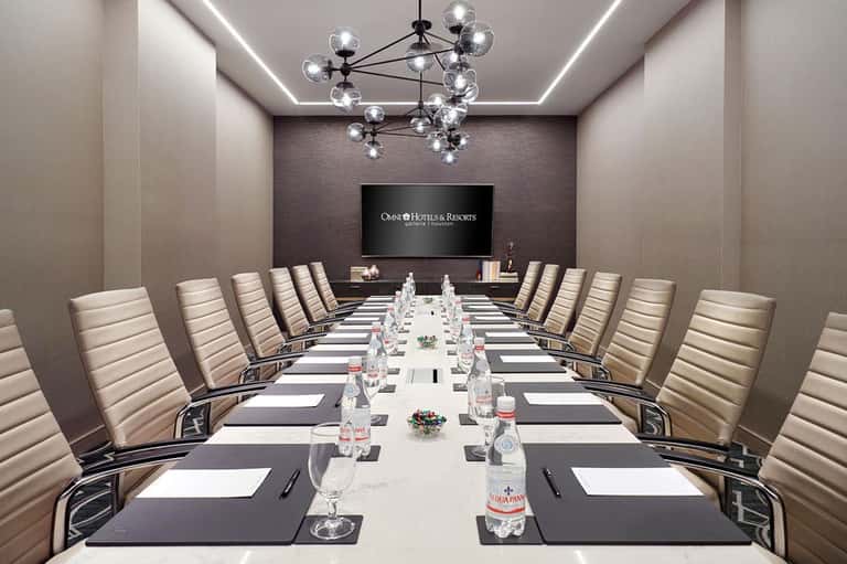 Modern conference room with long table, beige chairs, chandelier, and branded screen display