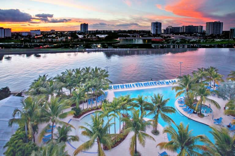 Resort pool complex at sunset with palm trees, multiple pools, and waterfront cityscape