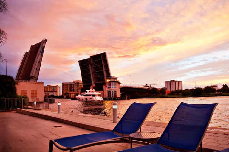 Waterfront sunset view with blue lounge chairs, drawbridge, and yacht on intracoastal waterway