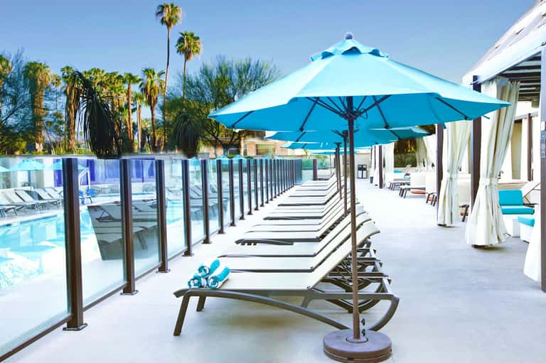 Poolside lounge area with blue umbrellas, loungers, and palm trees under clear sky