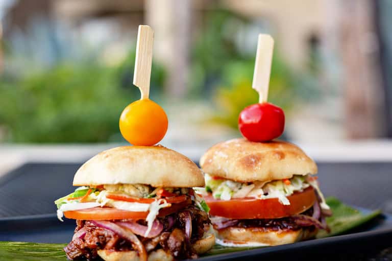 Two gourmet sliders with pulled meat, topped with colorful cherry tomatoes on wooden skewers, served on a dark plate