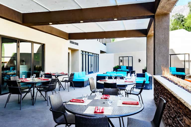 Modern courtyard dining area with round tables, blue cushioned seating, and covered pergola with fireplace