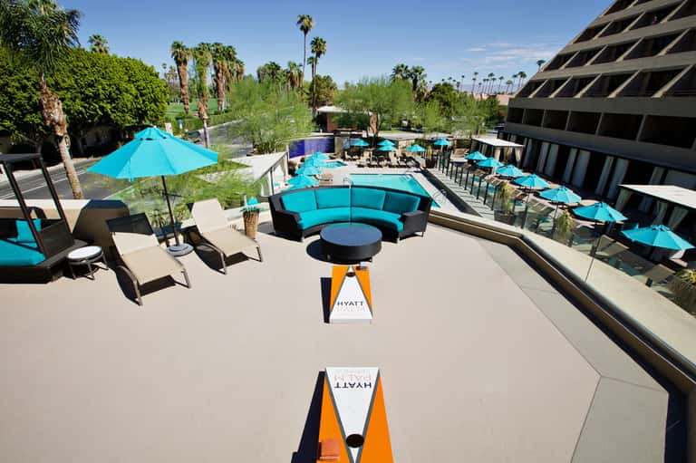 Resort pool area with bright turquoise umbrellas, curved seating, and desert landscaping with palm trees