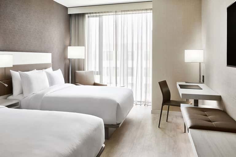 Modern hotel room with two queen beds, work desk, and floor-to-ceiling windows with sheer curtains