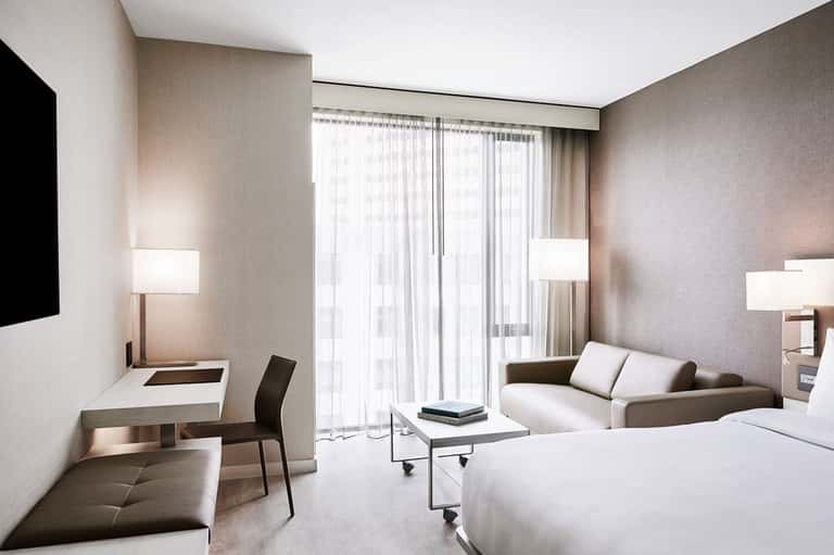 Modern hotel room with neutral tones, queen bed, work desk, sofa, and floor-to-ceiling windows with sheer curtains