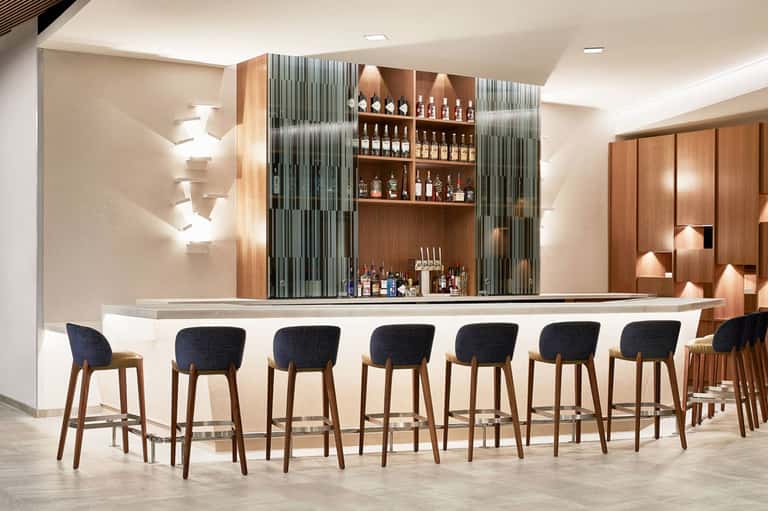 Modern hotel bar with curved white counter, wooden back shelving with bottles, and dark upholstered bar stools with wooden legs