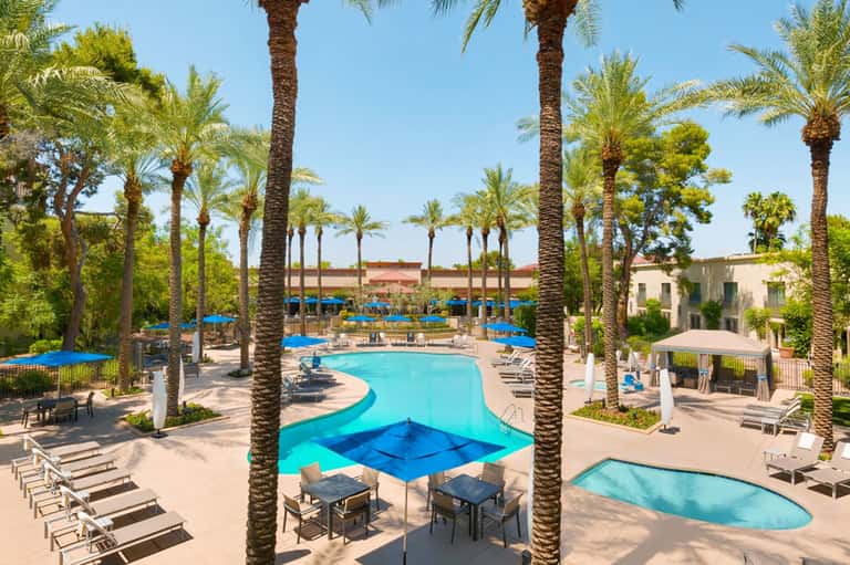 Resort pool area with turquoise water, palm trees, blue loungers, and beige umbrellas under clear sky
