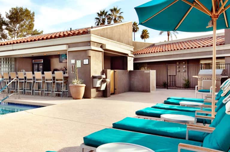 Resort poolside with turquoise lounge chairs, teal umbrella, and Spanish tile-roof buildings with palm trees