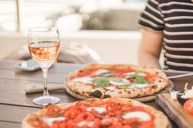 Rose wine glass and Margherita pizzas on outdoor dining table with striped shirt visible