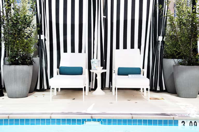 Two white lounge chairs under striped black and white canopy by poolside
