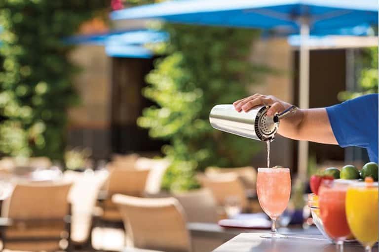 Bartender pouring cocktail at outdoor poolside bar with lounge chairs and blue umbrella