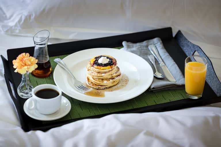 In-room breakfast tray with stacked pancakes, coffee, orange juice, and fresh flowers on a bed