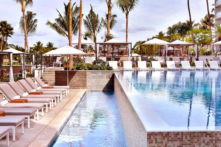 Luxury resort pool deck with palm trees, white lounge chairs, umbrella shades, and landscaping