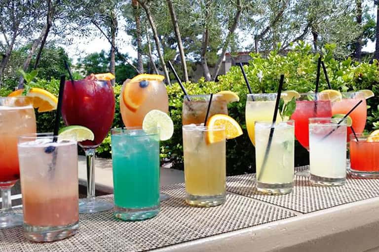 Array of colorful cocktails with fruit garnishes displayed outdoors on a patterned table with trees in background