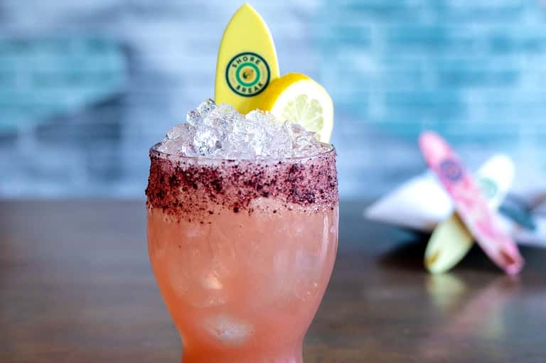 Colorful cocktail with pink liquid, crushed ice rim, and yellow surfboard garnish by ocean