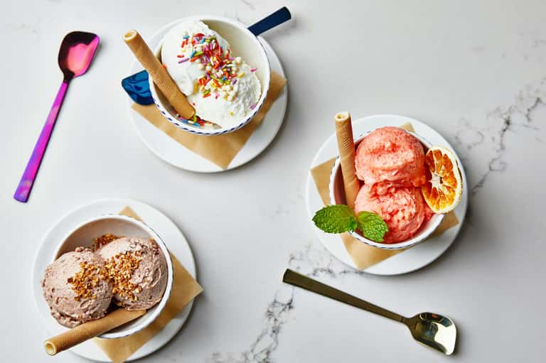Three ice cream bowls with colorful sprinkles, chocolate, and grapefruit garnish on marble surface