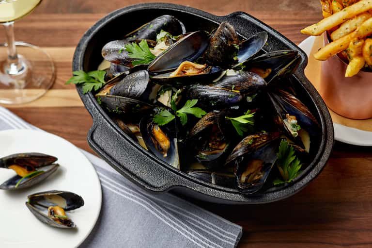 Black cast iron bowl of steamed mussels with parsley garnish, fries, and white wine on wooden table