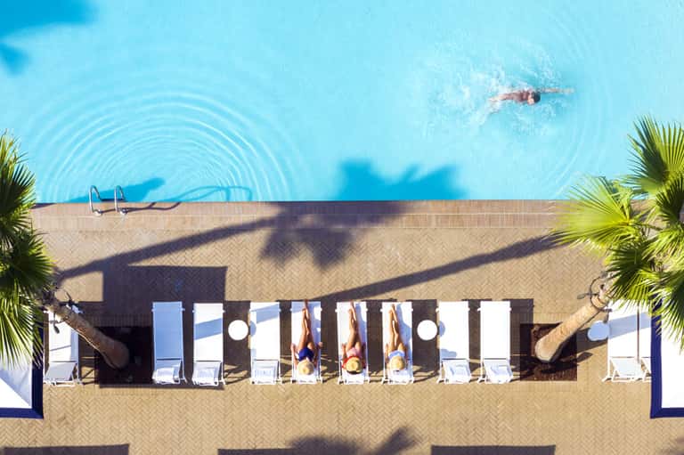 Overhead view of swimmer in bright turquoise pool with palm tree shadows and poolside loungers