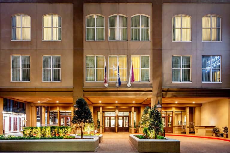 Hotel entrance at dusk with illuminated facade, large windows, and decorative planters flanking glass doors.