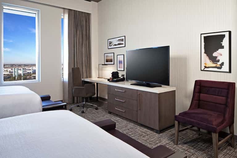 Modern hotel room with two beds, desk with TV, chair, and floor-to-ceiling window with city view