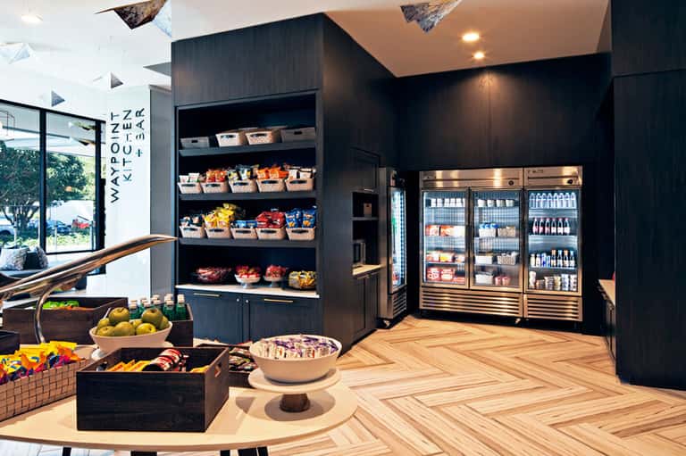 Modern resort marketplace with dark wood shelving, refrigerated beverage displays, fresh produce baskets, and herringbone wooden flooring