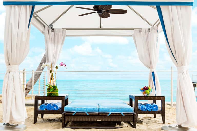 Luxury beach cabana with white curtains, ceiling fan, blue cushioned seating, ocean view, and rolled towels