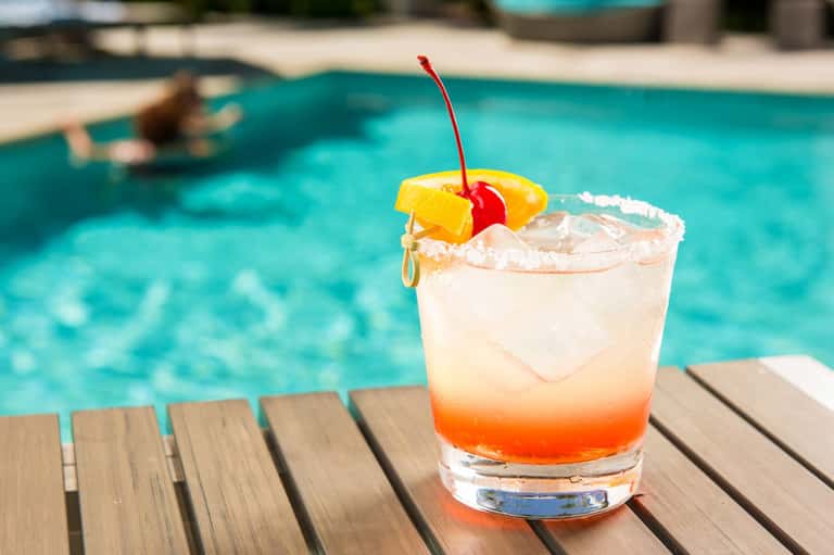Colorful layered cocktail with cherry and pineapple garnish on poolside deck overlooking blue pool