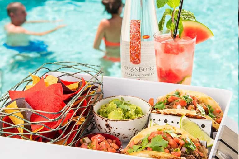 Poolside food and beverages including tacos, guacamole, colorful cocktail, and tortilla chips with vodka bottle