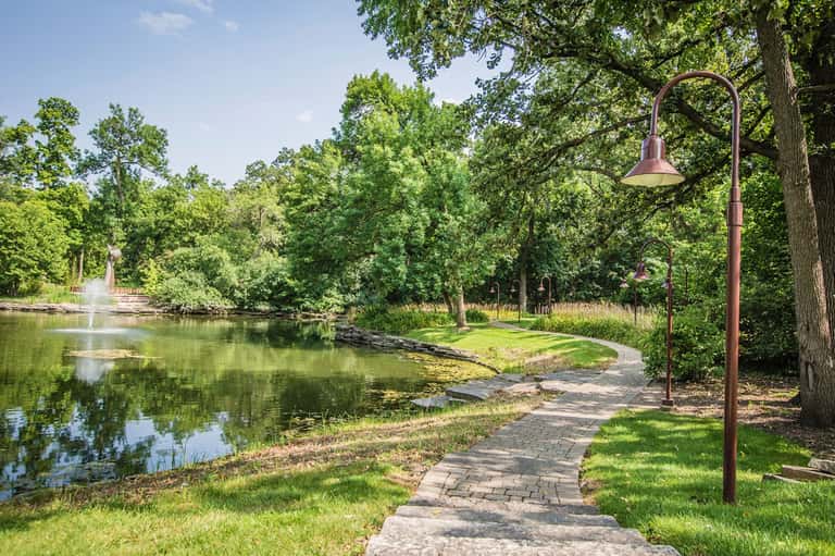 Scenic paved pathway along a calm pond surrounded by lush green trees and landscaping with a vintage street lamp