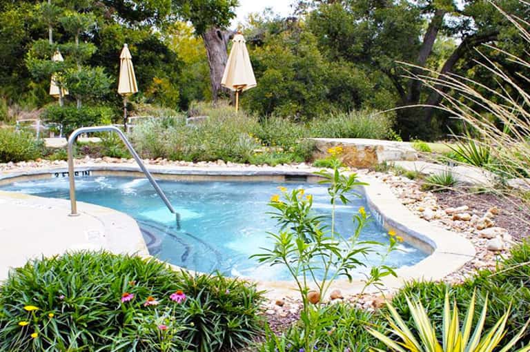 Resort pool with blue water surrounded by lounge chairs, umbrellas, and lush tropical landscaping with mature trees
