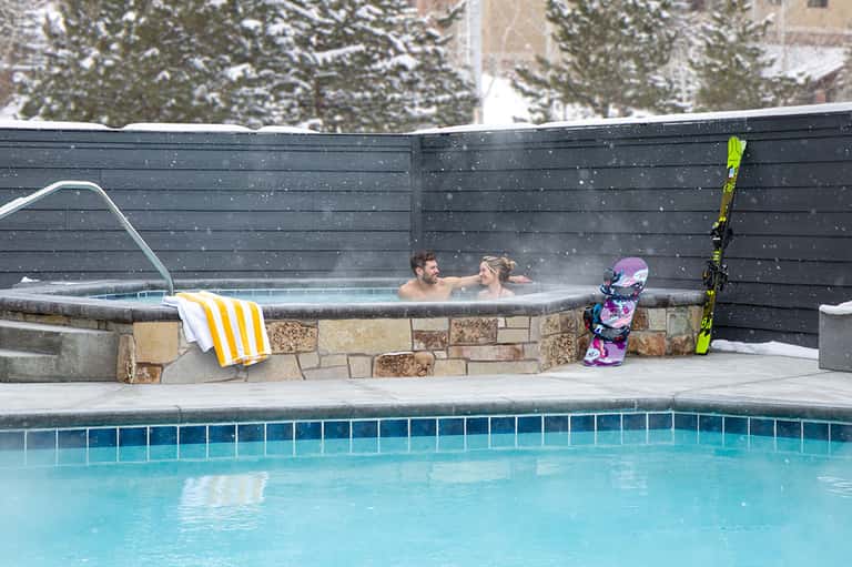 Couple relaxing in hot tub outdoors during winter snowfall with ski equipment nearby