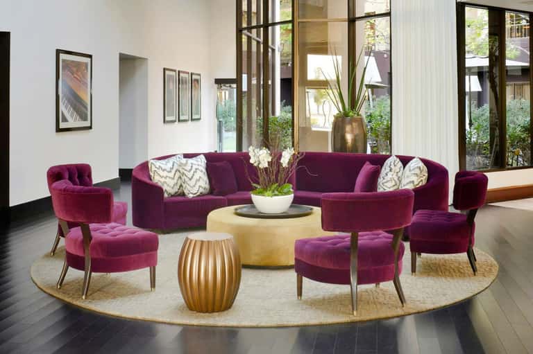 Modern lounge with purple velvet seating, round coffee tables, and large windows overlooking a courtyard garden.