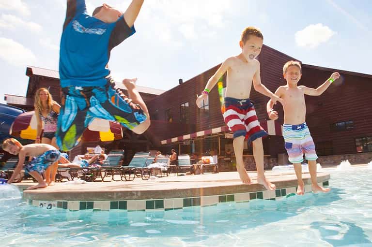 Children jumping into a resort pool on a sunny day with lounge chairs and brown building in background