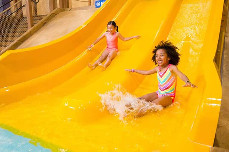Two children sliding down a bright yellow water slide with water splash at an indoor aquatic center