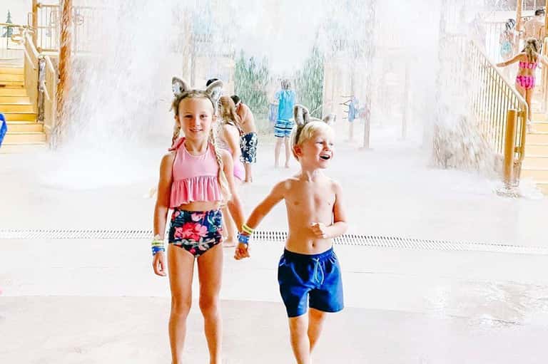 Children running through indoor water park splash zone with white walls and yellow railings