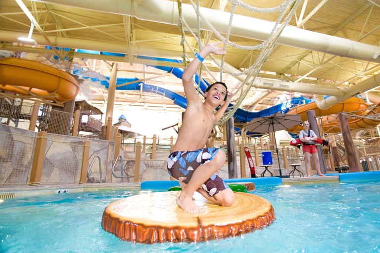 Young boy on inflatable platform in indoor water park with rope obstacles and slides
