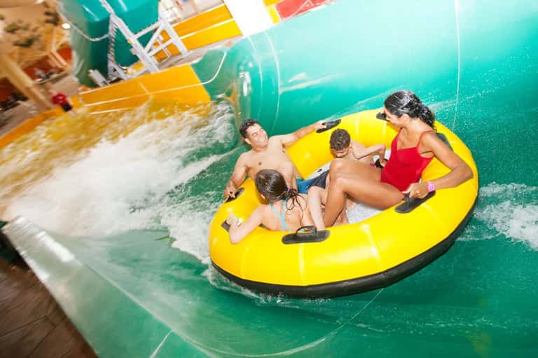Four guests on yellow inflatable raft riding water slide into turquoise pool at resort