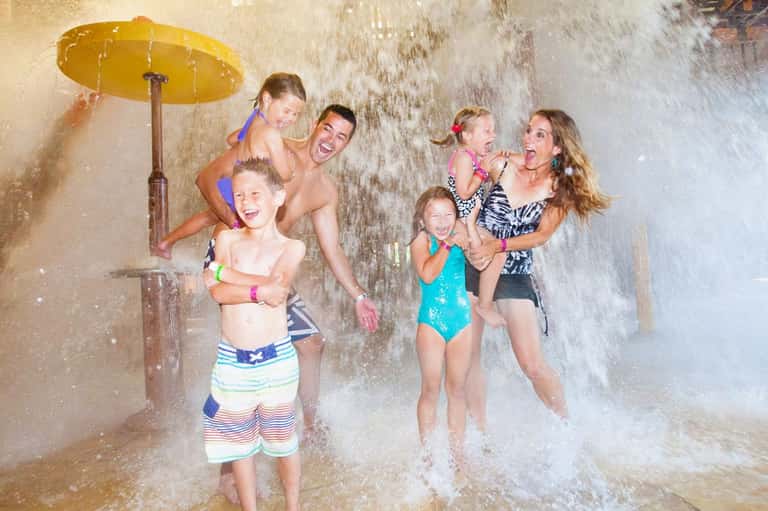Family enjoying water play under an outdoor shower with yellow umbrella, children and adults splashing together