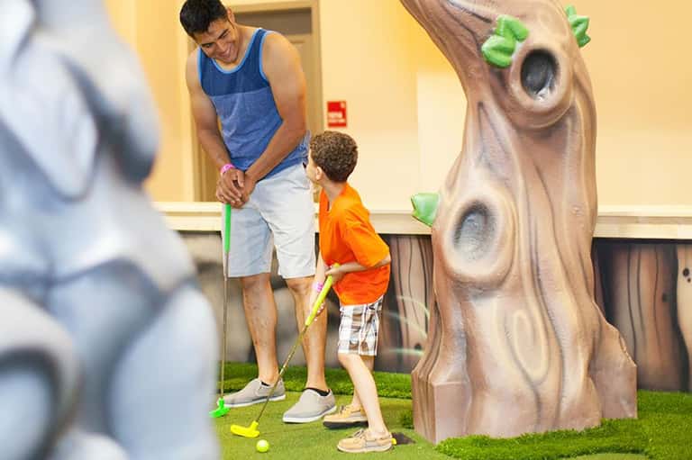 Man and child playing mini golf indoors with tree-themed obstacles and green carpet