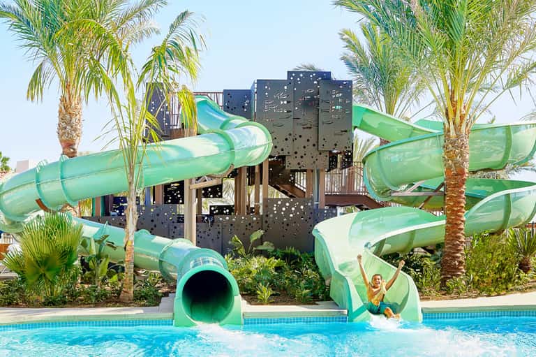 Green water slides at resort pool with palm trees and children playing in turquoise water
