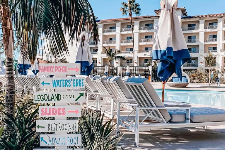 Resort pool with directional signs to family pool, water slide, and adult pool, blue umbrellas, striped lounge chairs, palm trees