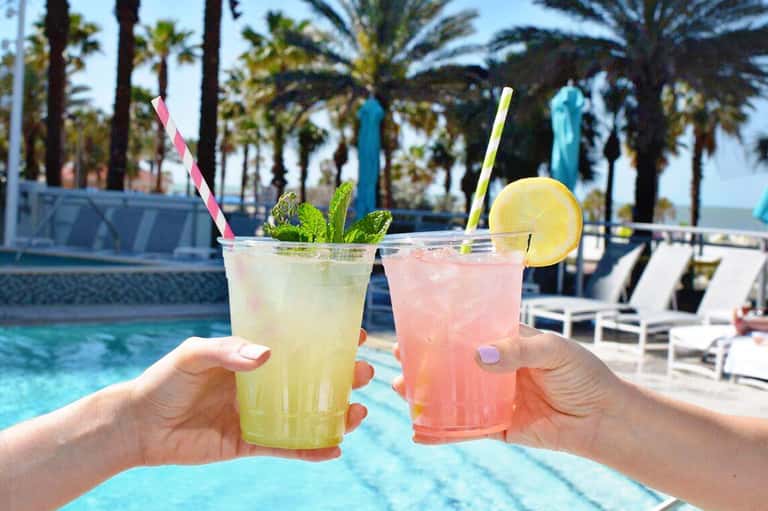 Two colorful cocktails with fresh garnishes by a resort pool and palm trees