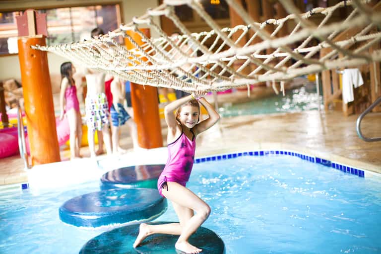 Young girl in pink swimsuit playing in indoor resort pool with rope bridge and colorful columns