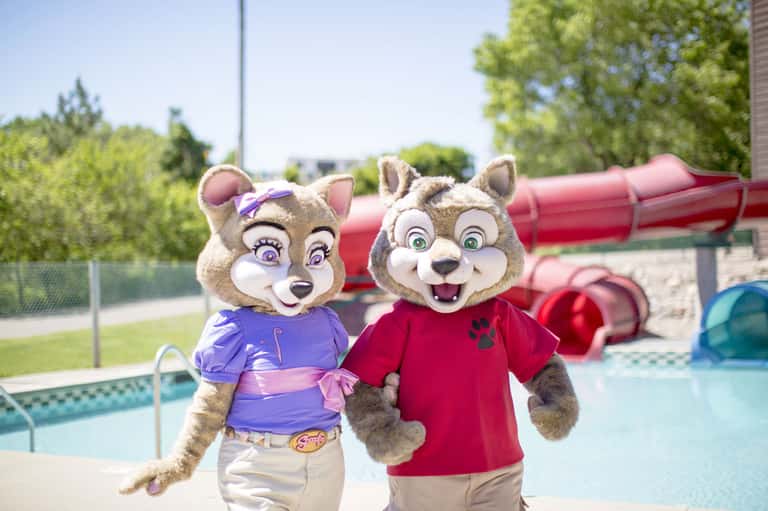 Mascot characters in purple and red outfits posing by a resort pool with water slides