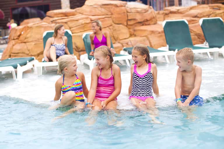 Children playing in resort pool with rocky features and adults relaxing on lounge chairs nearby