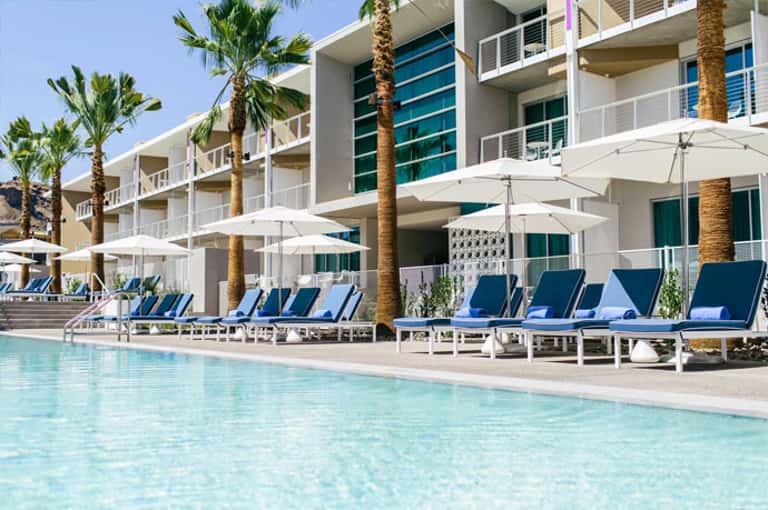 Modern resort pool with palm trees, white umbrellas, and blue loungers beside contemporary building