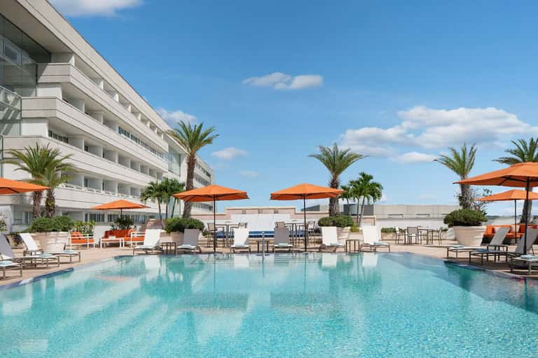 Large resort pool surrounded by palm trees, orange umbrellas, and modern hotel building