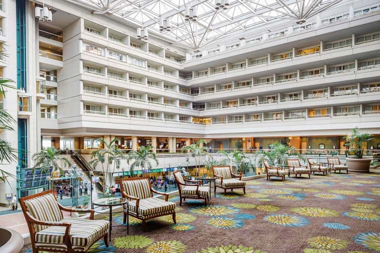 Multi-story hotel atrium with striped seating, tropical plants, patterned flooring, and natural skylight